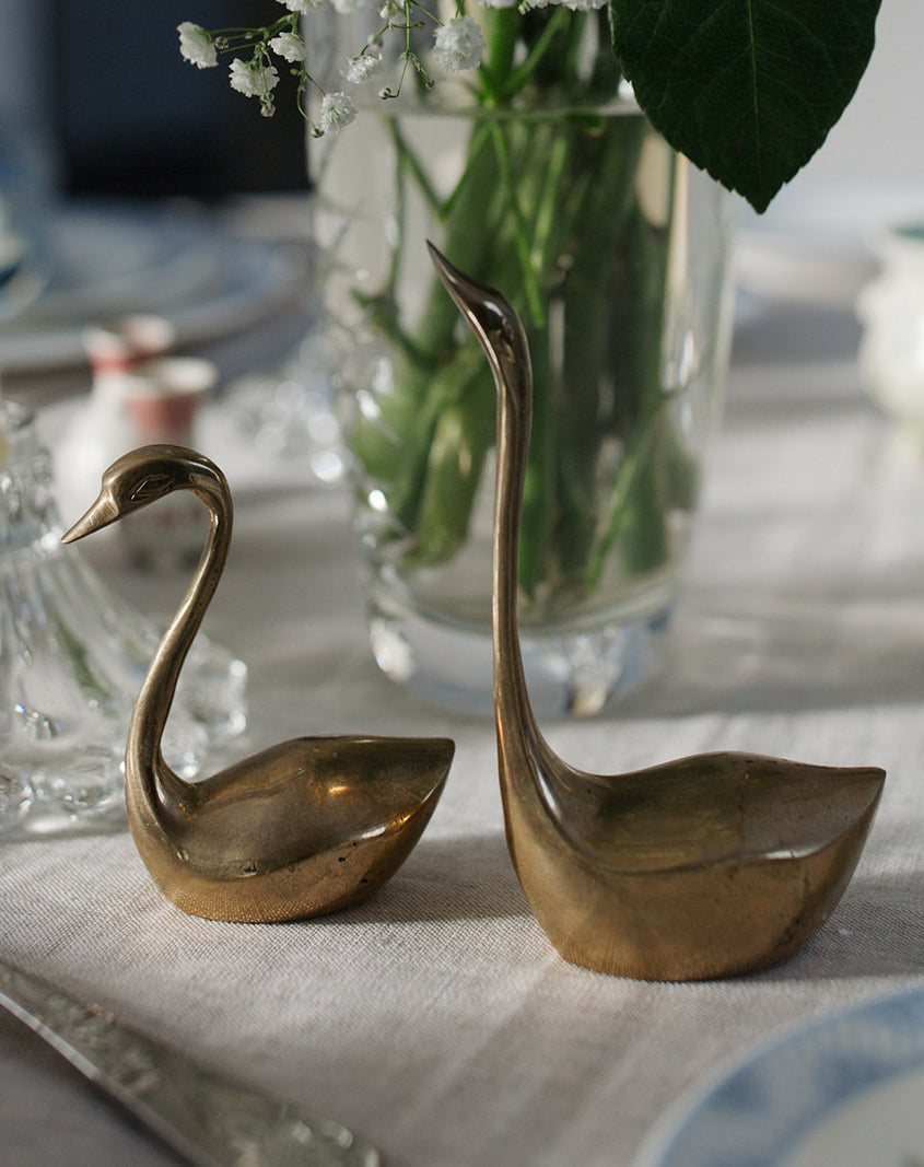 Two brass swans sitting on linen tablecloth in front of vase of florals and greenery.